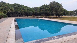 A rectangular swimming pool with a wooden deck surrounding it, set in a landscaped area with trees and a clear sky. - Mango Retreat Hotel and Resort (Bhopal)