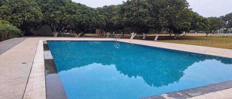 Piscina retangular com deck de madeira ao redor, situada em área ajardinada com árvores e céu claro.