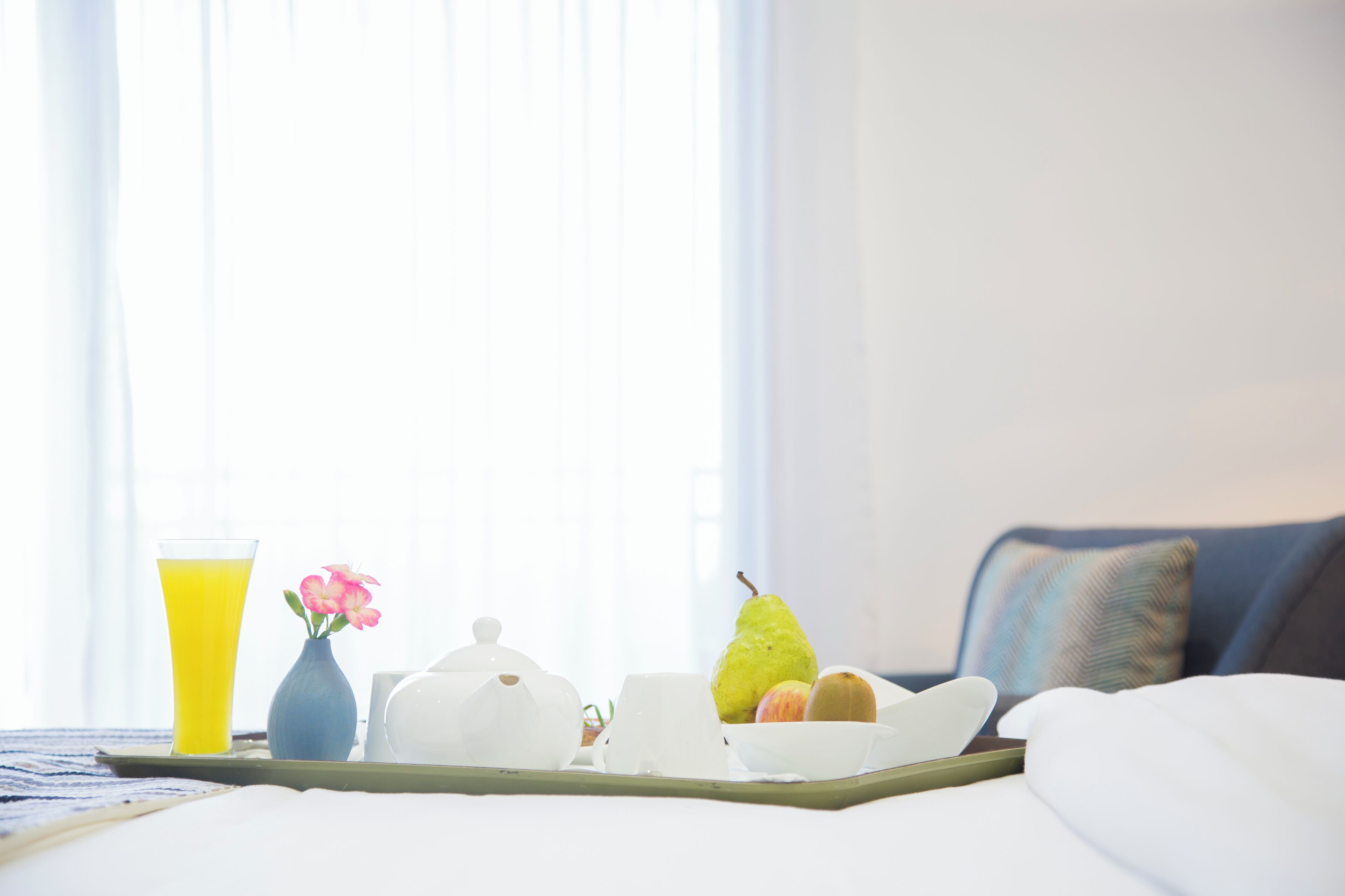 A breakfast tray with a glass of orange juice, a vase with pink flowers, a teapot, a pear, a pumpkin, and a bowl of eggs on a bed.