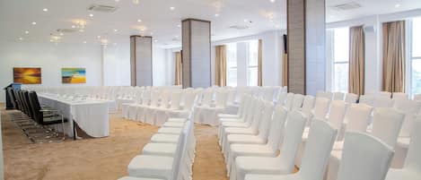 A conference room with rows of white chairs, a long table with chairs, and large windows with curtains.