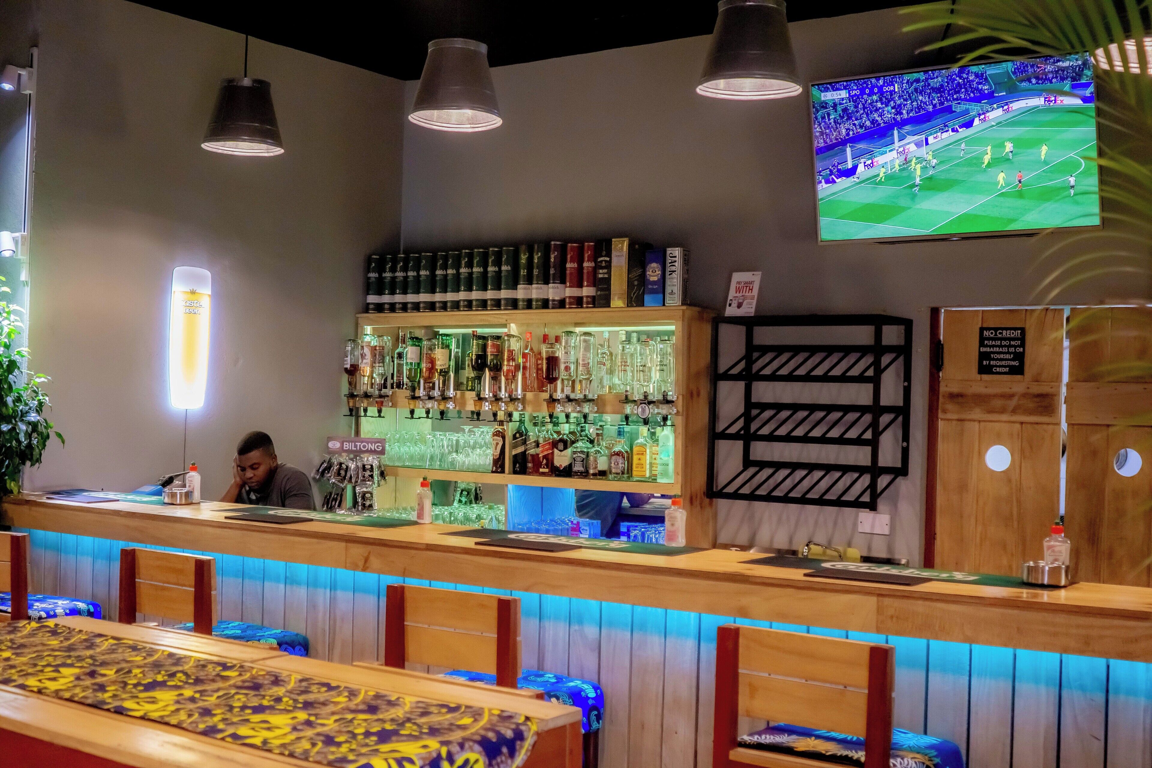 Un espace bar avec un comptoir, des étagères remplies de bouteilles d’alcool et un téléviseur diffusant un match de football.