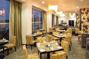 A hotel dining area with multiple tables and chairs, large windows, and modern lighting fixtures.