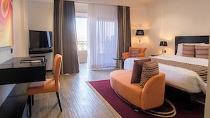 A hotel room with a bed, a desk with a TV, a sofa, and a balcony with curtains.