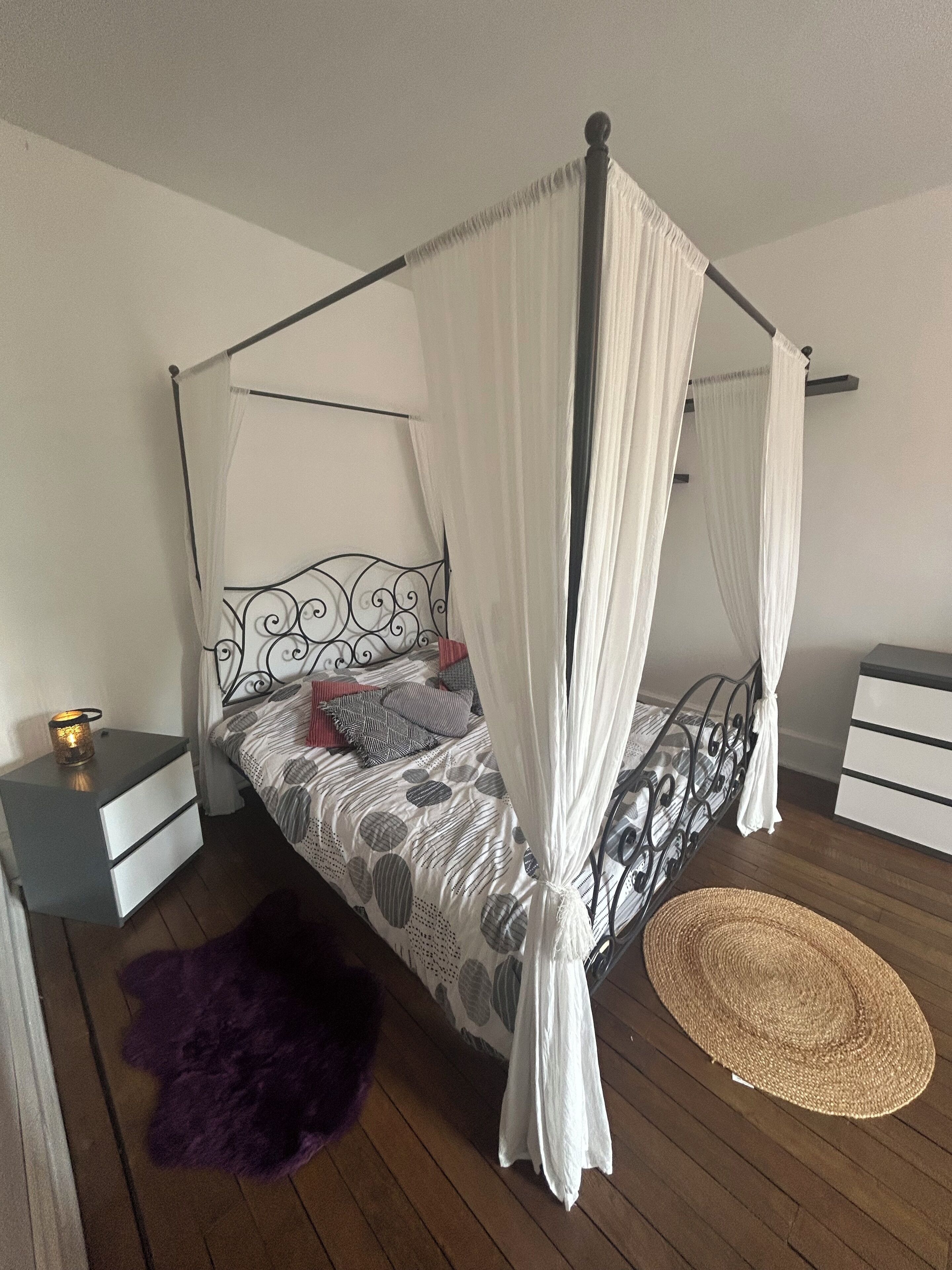 Una cama con dosel y cortinas blancas, un edredón estampado y una mesita de noche con lámpara.