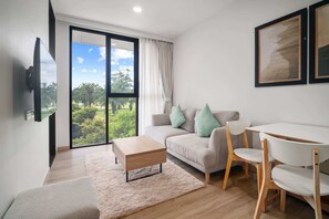 Un salon moderne comprenant un canapé, une table basse, une table à manger avec des chaises, un téléviseur fixé au mur et une grande porte-fenêtre coulissante donnant sur un balcon avec vue sur la verdure.