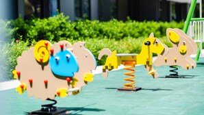 Des appareils de musculation en plein air, dotés d’un système à ressort, installés sur un fond de verdure et avec des bâtiments modernes en arrière-plan.