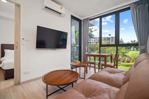 A modern hotel room with a large window, a flat-screen TV, a sofa, a small table, and a bed.