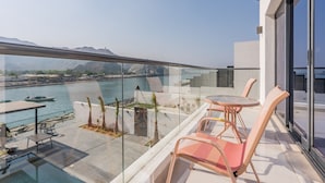 Ein Balkon mit Glastrennwand, der auf ein Gewässer und Berge blickt. Auf dem Balkon befinden sich ein Tisch und Stühle.