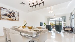 Ein moderner Essbereich mit einem weißen Tisch, gedeckt für sechs Personen, umgeben von weißen Stühlen. An der Wand hängt ein großes Gemälde eines Pferdes, über dem Tisch hängt eine Chandelüster und durch große Fenster ist die Natur zu sehen.