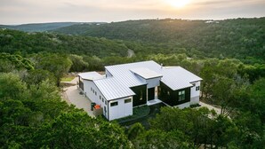 A modern two-story house with a flat roof, surrounded by dense forest. - Labor Day Getaway | Heated Pool + Spa + Epic Views (Canyon Lake)
