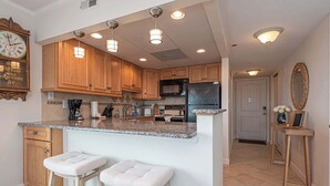 A kitchen with wooden cabinets, a granite countertop, two bar stools, a clock, and a refrigerator. - Ocean Front Condo with Pool (Ocean City)