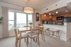 A kitchen with wooden cabinets, a dining table with chairs, a bar stool, a clock, and a window with curtains. - Ocean Front Condo with Pool (Ocean City)
