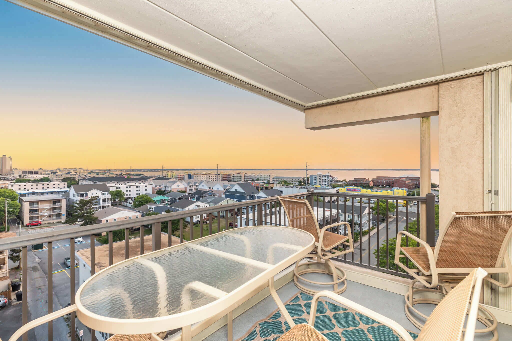 A balcony with a glass table and wicker chairs, overlooking a residential area with houses and a clear sky.