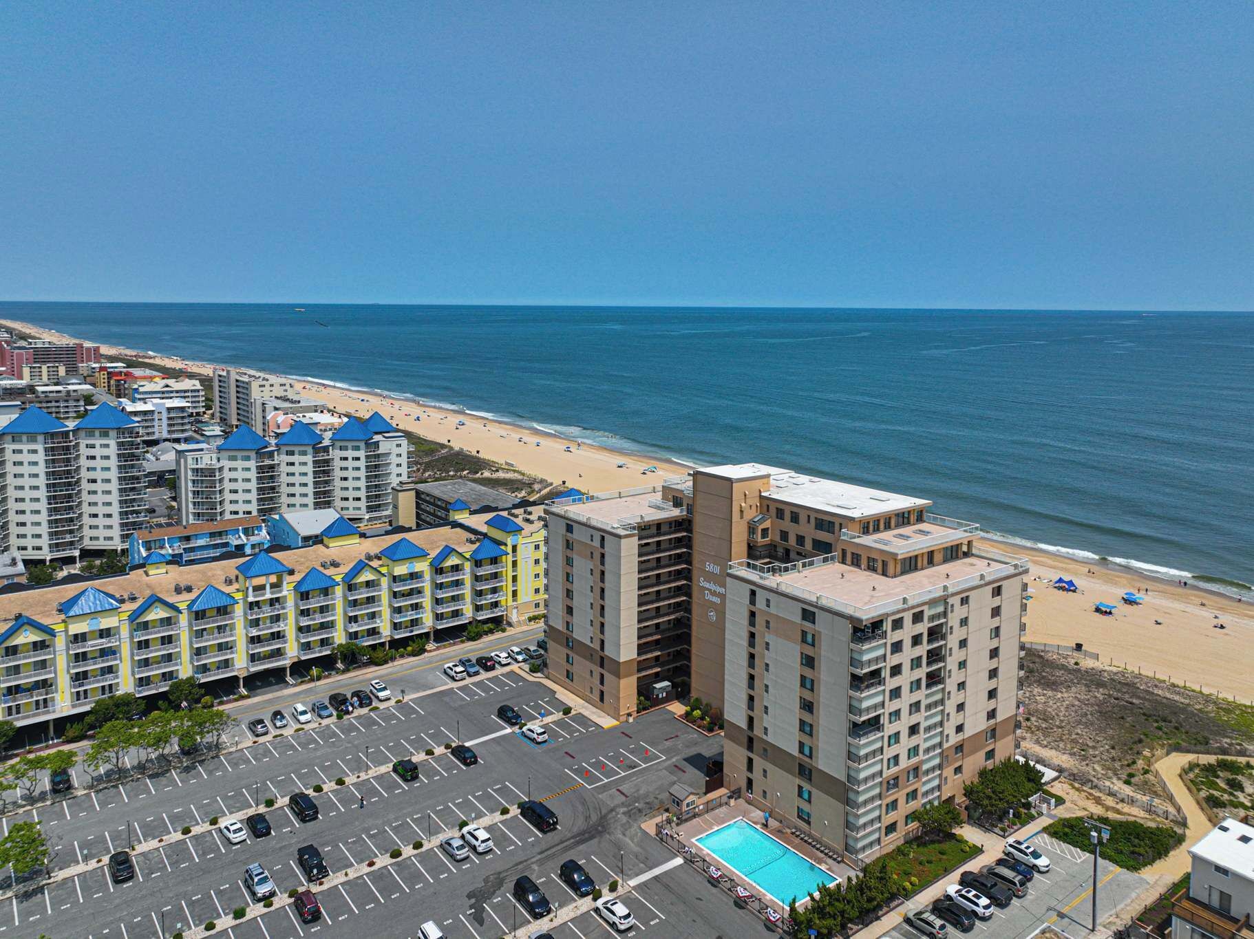 A coastal hotel with multiple buildings, a parking lot, and a beach view.