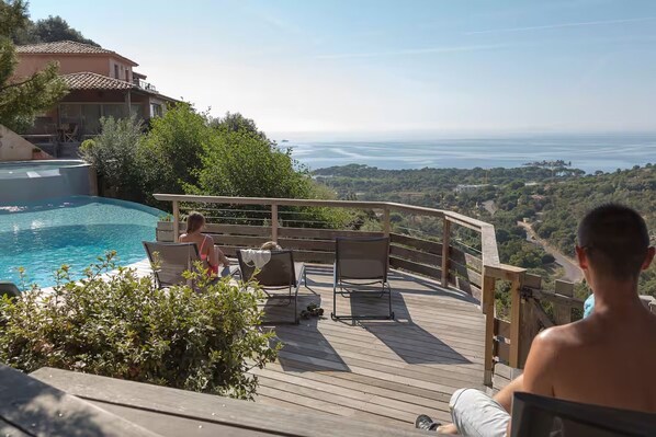 A poolside area with a wooden deck, lounge chairs, and a view of the sea.