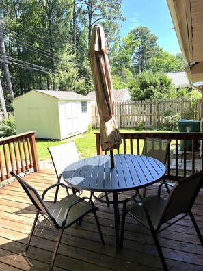 A wooden deck with a round table and chairs, an umbrella, and a white picket fence in the background.