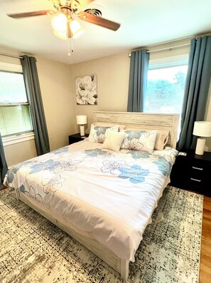 A neatly made bed with a floral bedspread, a ceiling fan, and a window with curtains.