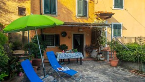 Un patio con suelo de piedra, dos sillones azules, un parasol verde y una mesa con mantel blanco.