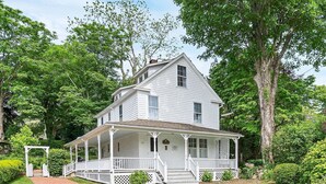 A white two-story house with a porch, surrounded by lush greenery.