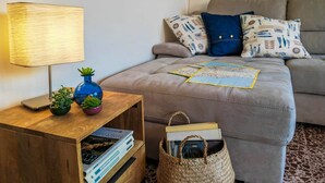 Un salon chaleureux avec un canapé, une lampe, une petite table d’appoint en bois sur laquelle sont posés des livres et une plante, et un panier en osier.