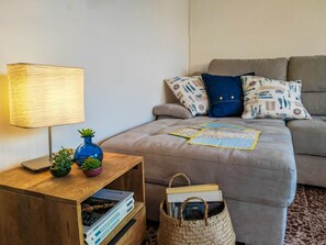 A cozy living room with a sofa, a lamp, a small wooden side table with books and a plant, and a woven basket.