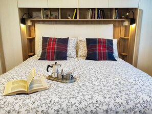 Uma cama bem arrumada com um cobre-leito floral, duas almofadas xadrez, uma mesa de cabeceira com um livro e uma cafeteira, e uma prateleira com livros e uma pequena planta.