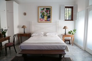 Een slaapkamer met een bed, nachtkastjes, een stoel, een bureau, een lamp, een potplant en een raam met luiken.