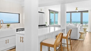 A modern kitchen with white cabinetry, a central island with bar stools, and a view of the ocean through large windows. - Rare Front Stack Remodeled 2 bedroom Gem in Mainsail- Free Beach Service! (Miramar Beach)