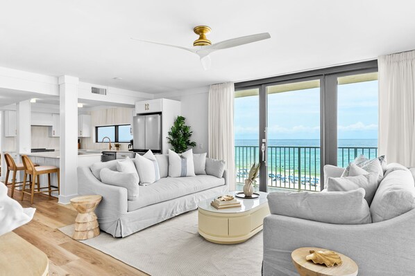 A modern living room with a sofa, a coffee table, and a dining area with chairs. There is a large window with a view of the ocean.