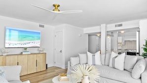 A modern living room with a large flat-screen TV, a white sofa, a coffee table with decorative items, and a wooden cabinet. - Rare Front Stack Remodeled 2 bedroom Gem in Mainsail- Free Beach Service! (Miramar Beach)