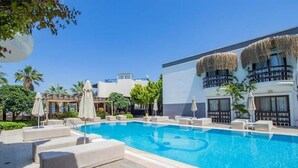 Una piscina con sillones de descanso y sombrillas, rodeada de vegetación y un edificio con fachada blanca y balcones de techo de paja.