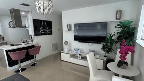 Una sala de estar moderna con un televisor de pantalla plana, un comedor con taburetes rosados, un sofá gris y una mesa de centro blanca.
