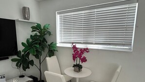 Un salón moderno con un televisor de pantalla plana, un sofá blanco, una mesita redonda con un jarrón de flores y un amplio ventanal con persianas.