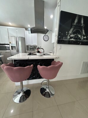 A modern kitchen with white cabinets, stainless steel appliances, a central island with pink bar stools, a clock, and a large black and white photo of the Eiffel Tower on the wall.