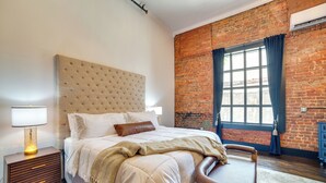 A bedroom with a large bed, a nightstand, a chair, and a brick wall. - Heart of Downtown Marshall: Boutique Studio Stay! (Marshall)