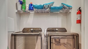 A laundry area with two stainless steel front-loading washing machines, a shelf above with cleaning supplies, and a fire extinguisher on the wall.