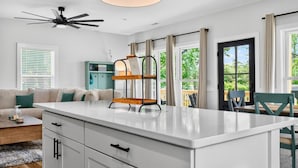 A modern kitchen with a central island, white cabinetry, and a ceiling fan. - Lux Oak Island Retreat | Walk to Beach | Private Suite + Fenced Yard, Swim Spa Coming Soon! (Oak Island)