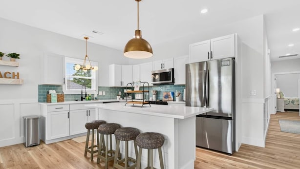 A modern kitchen with white cabinetry, stainless steel appliances, and a central island with bar stools. - Lux Oak Island Retreat | Walk to Beach | Private Suite + Fenced Yard, Swim Spa Coming Soon! (Oak Island)
