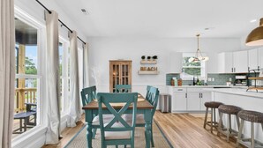 A modern kitchen with white cabinets, a wooden dining table with teal chairs, and a view of the outdoors through large windows. - Lux Oak Island Retreat | Walk to Beach | Private Suite + Fenced Yard, Swim Spa Coming Soon! (Oak Island)