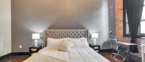 Un dormitorio con cabecera capitonada, una cama grande con ropa de cama blanca, un escritorio con silla y una pared de ladrillo.