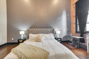 Une chambre avec une tête de lit capitonnée, un grand lit avec une literie blanche, un bureau avec une chaise et un mur en briques.