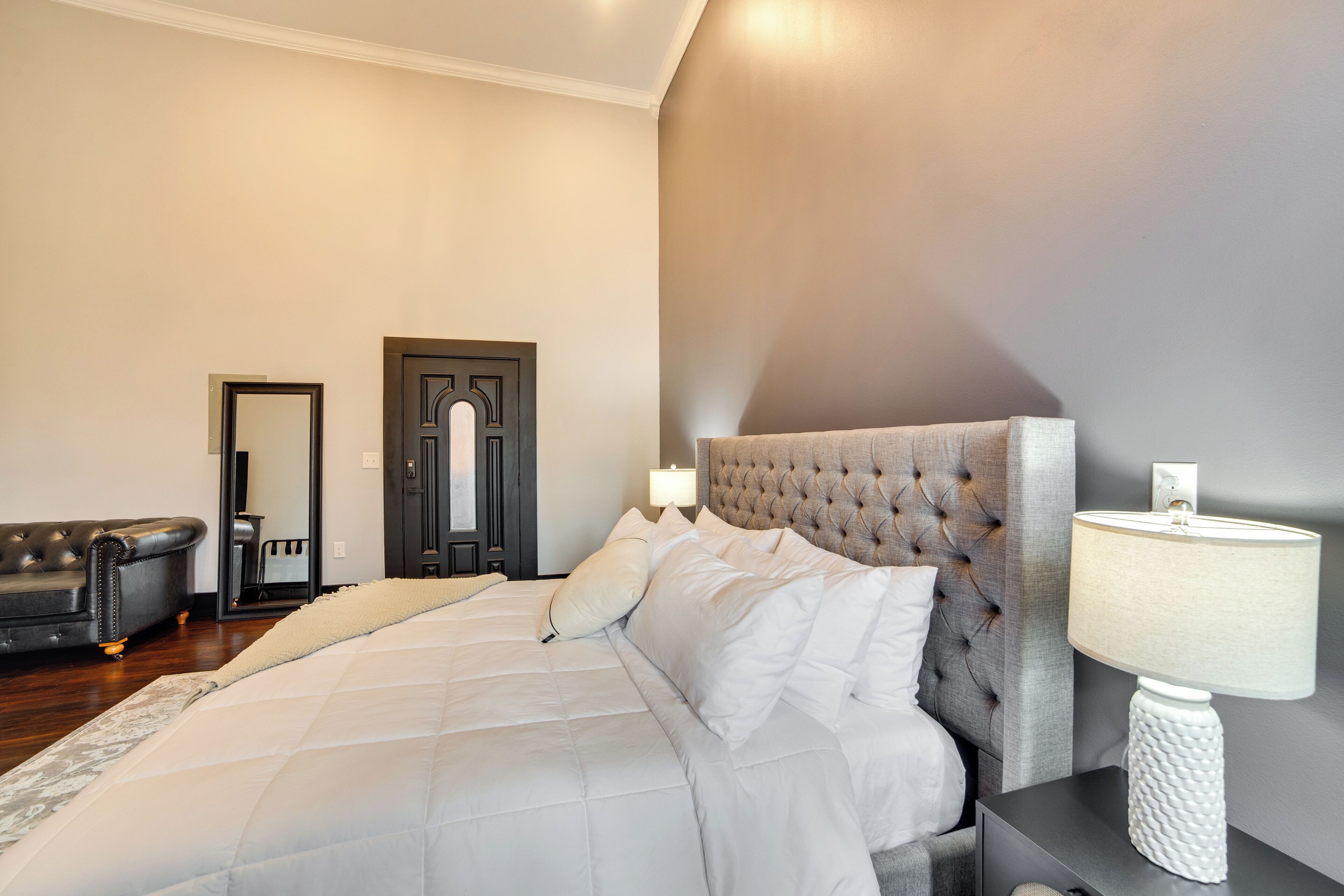 Un dormitorio con una cama grande, una mesita de noche con lámpara, una puerta de color oscuro y un sofá tapizado en gris.
