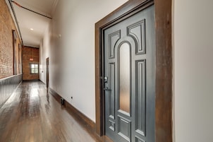 Un pasillo con piso de madera, una puerta oscura con picaporte y una pared de ladrillo en un lateral.