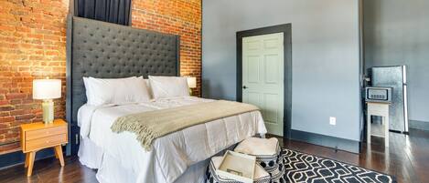Un dormitorio con pared de ladrillo, cabecera capitonada, cama blanca, lámparas de noche, alfombra estampada y cestas de almacenamiento.