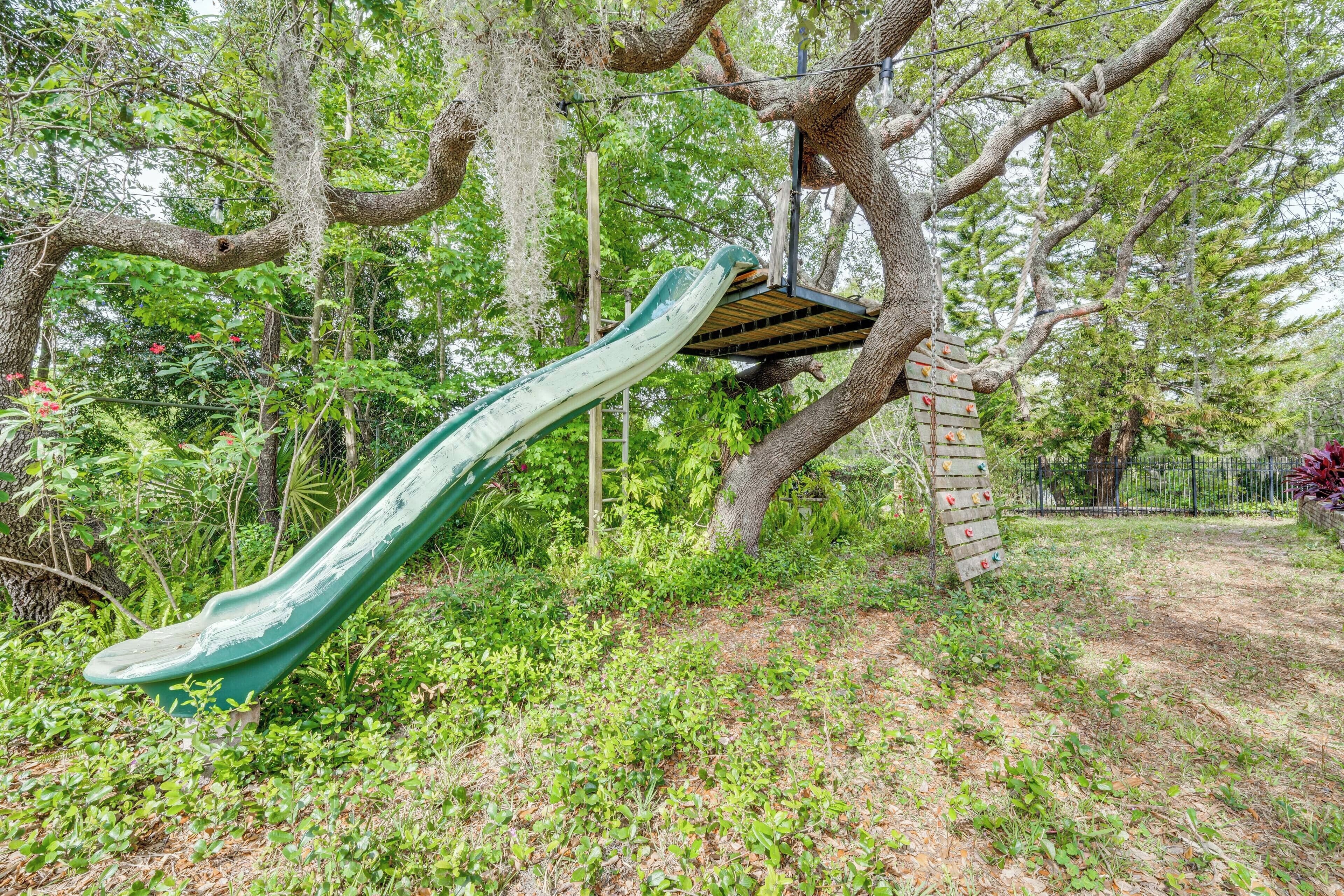 Un parque infantil con un tobogán verde y una estructura para escalar, rodeado de árboles y follaje.