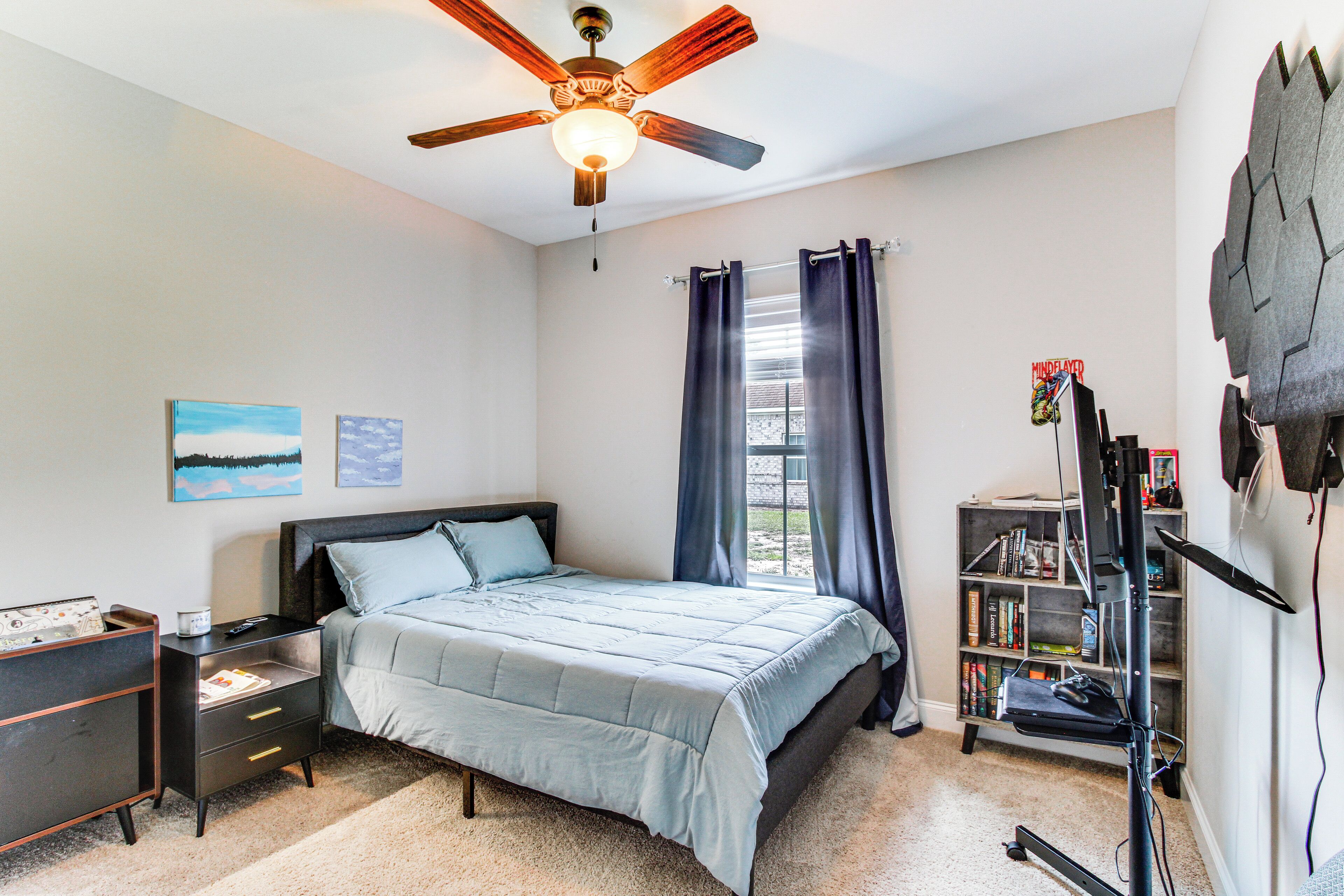 A bedroom with a bed, a nightstand, a bookshelf, a ceiling fan, and a window with curtains.