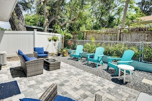 An outdoor patio area with wicker furniture, including chairs and a table, surrounded by a white fence and greenery.