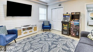 A living room with a flat-screen TV, a blue armchair, a grey sofa, a patterned rug, and arcade games. - Apt 3 Mi to Beach & Daytona Boardwalk Amusements! (Holly Hill)