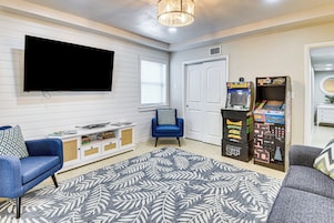 A living room with a flat-screen TV, a blue armchair, a grey sofa, a patterned rug, and arcade games.