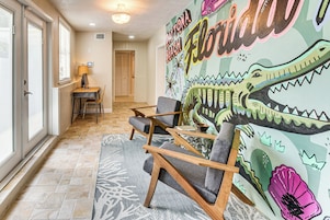 Una habitación con un gran mural en la pared, una silla de madera, una mesita y una lámpara.